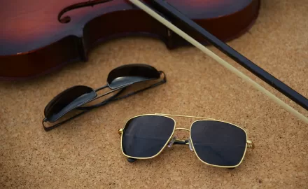 Óculos de sol aviador dourado e preto sobre superfície de cortiça, ao lado de violino